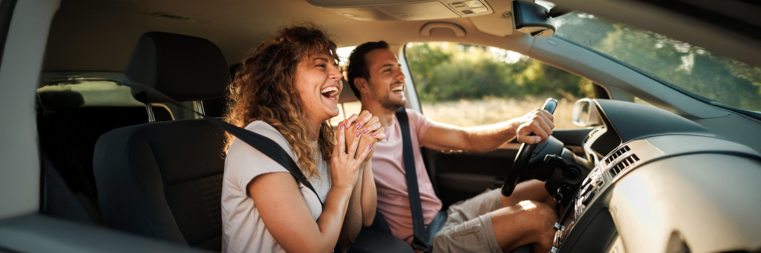 Two people in a car, one driving, with greenery outside.