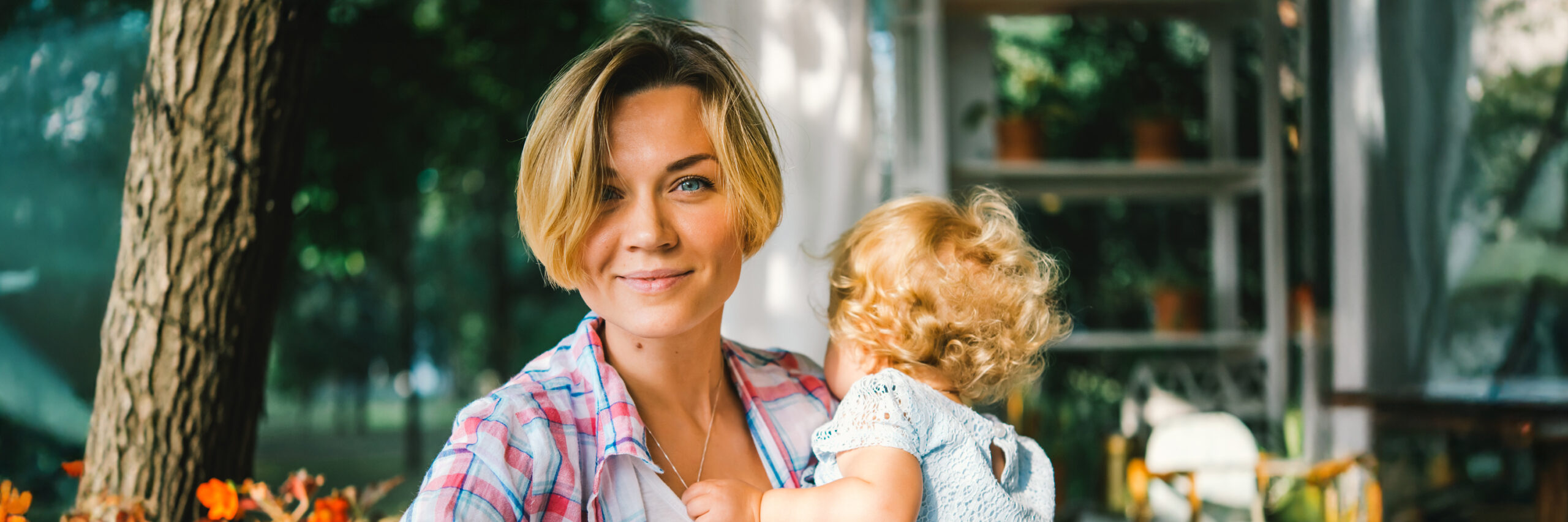 A person holding a child outdoors near plants and a seating area.