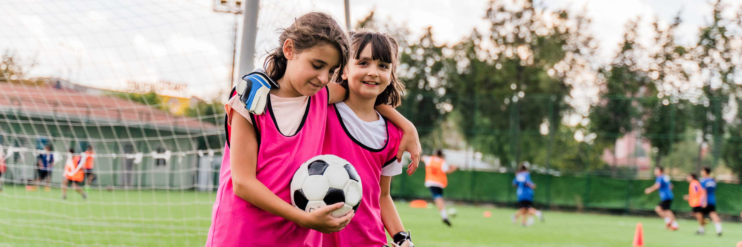 Two children in pink vests on a soccer field, one holding a ball, with others playing in the background.