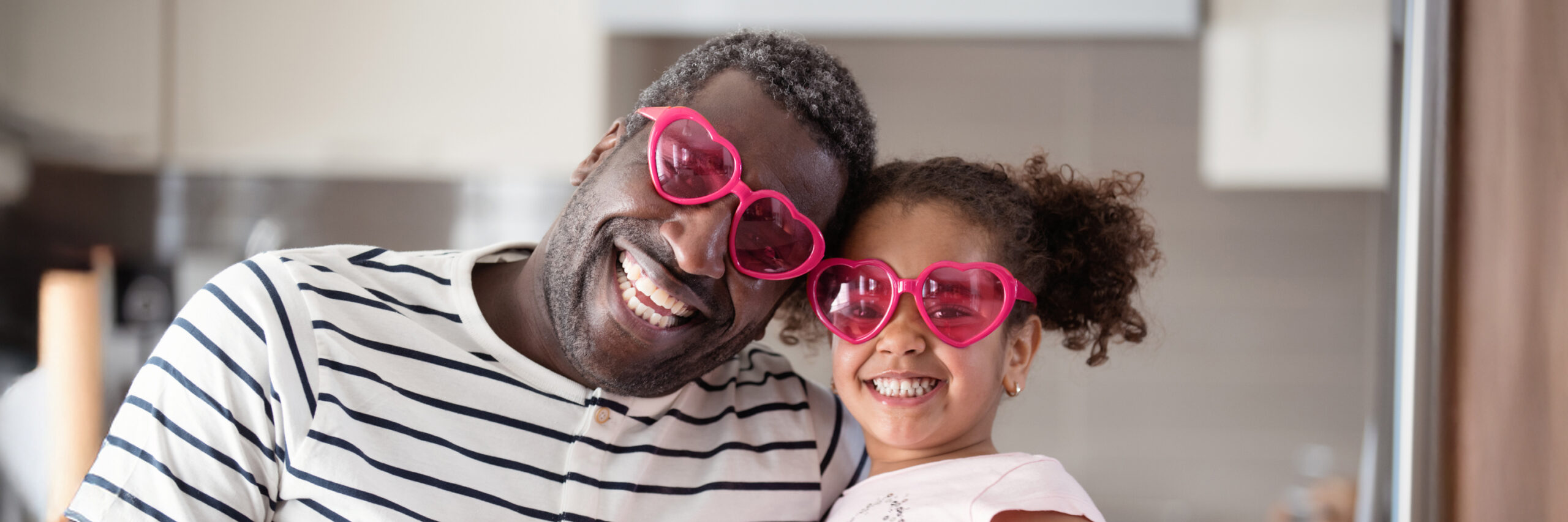 A parent and child wearing heart-shaped glasses, sitting together and smiling indoors.