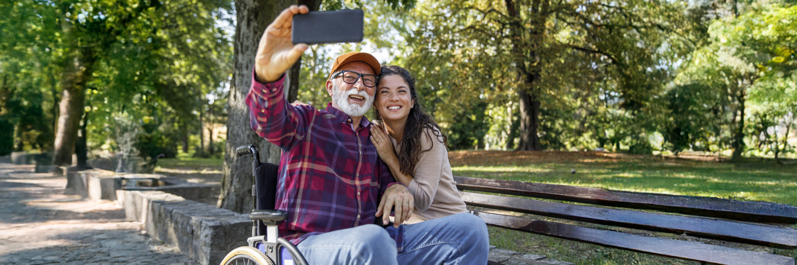 Two people taking a selfie on a park bench, one in a wheelchair.