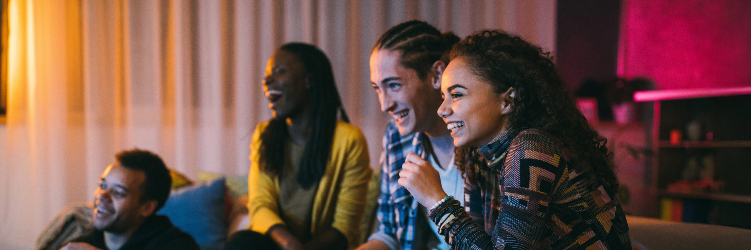 Four people smiling and sitting together in a dimly lit room with curtains and shelves in the background.