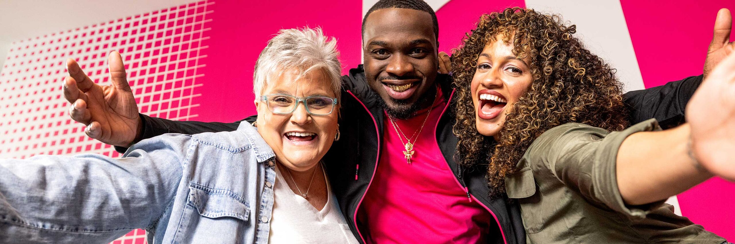 Three T-Mobile employees smiling at the camera in front of a bright pink T-Mobile branded wall.
