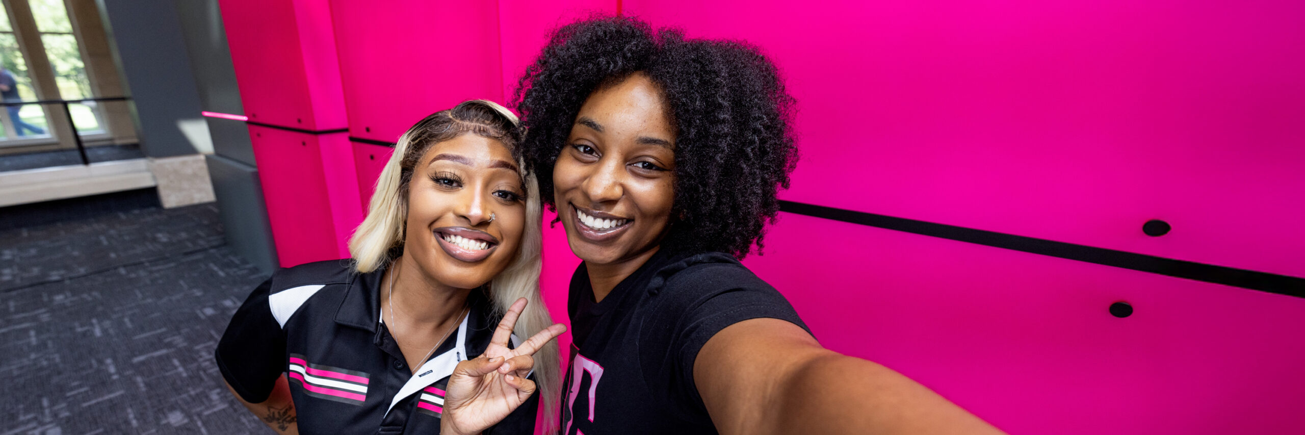 Two T-Mobile employees taking a selfie in front of a bright pink wall, one making a peace sign.