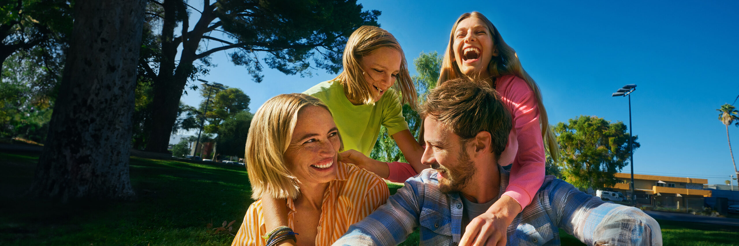 A family laughing outdoors in a park with trees and a blue sky.
