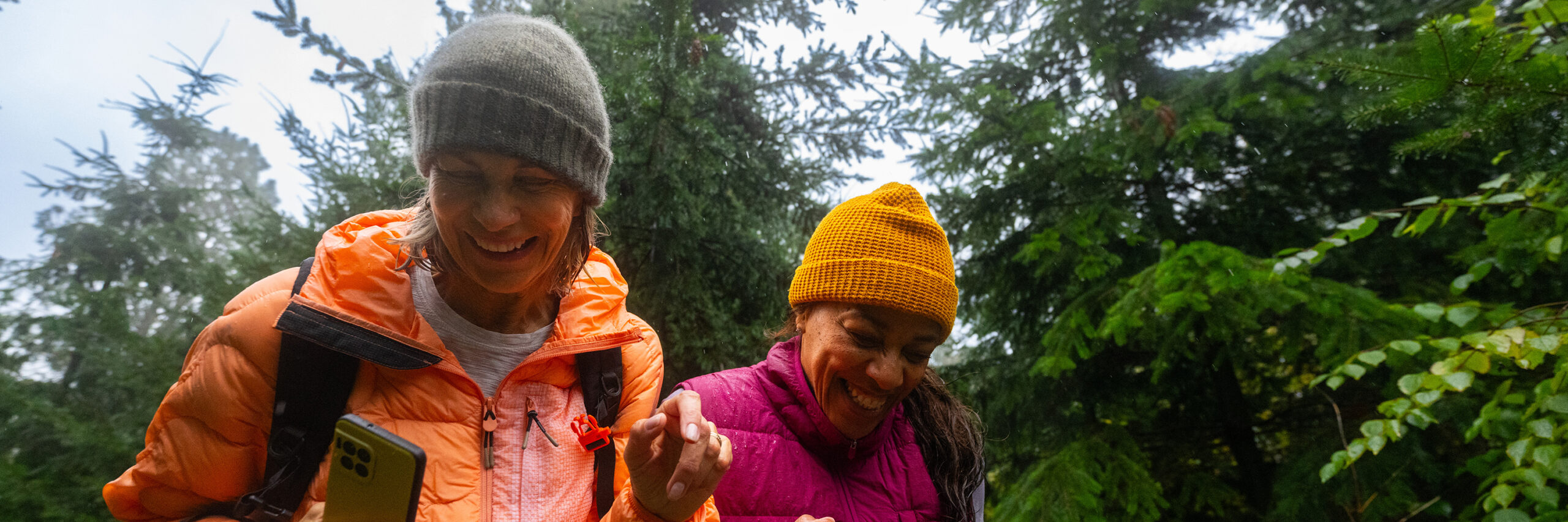 Two people smiling and laughing outdoors in a forested area, one holding a phone.