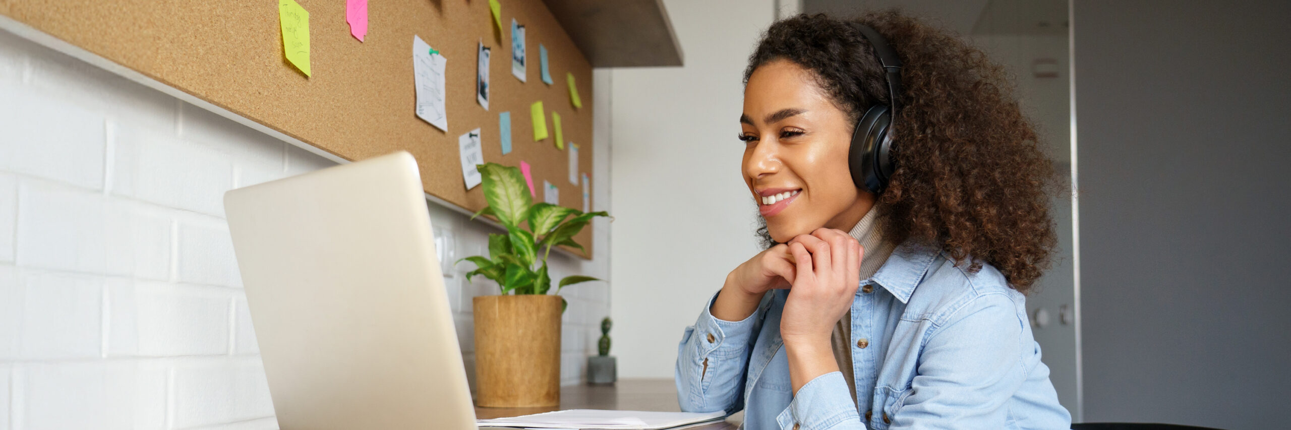 A student with headphones working on a laptop at a desk with a corkboard.