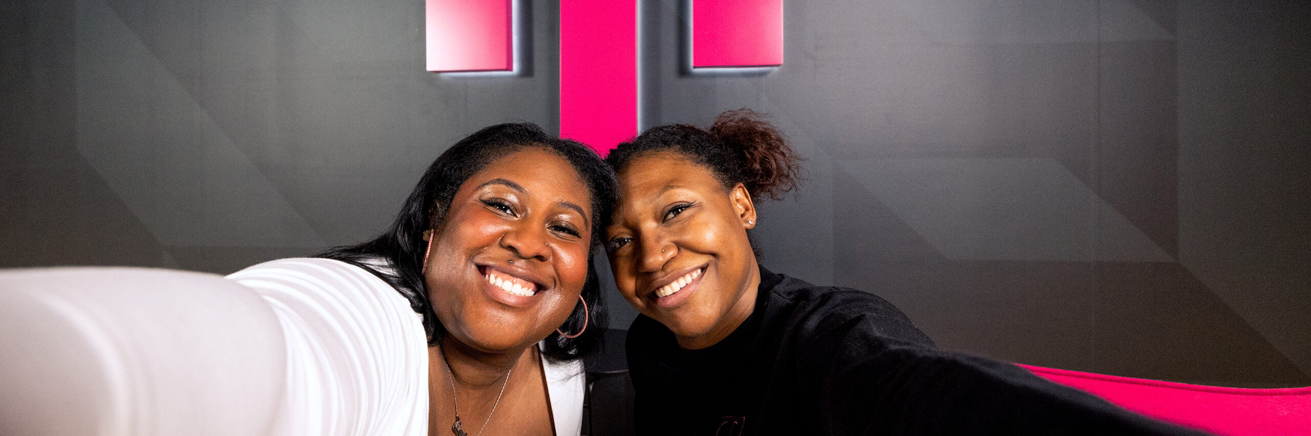 Two T-Mobile employees taking a selfie in front of a pink T-Mobile sign on a gray wall.