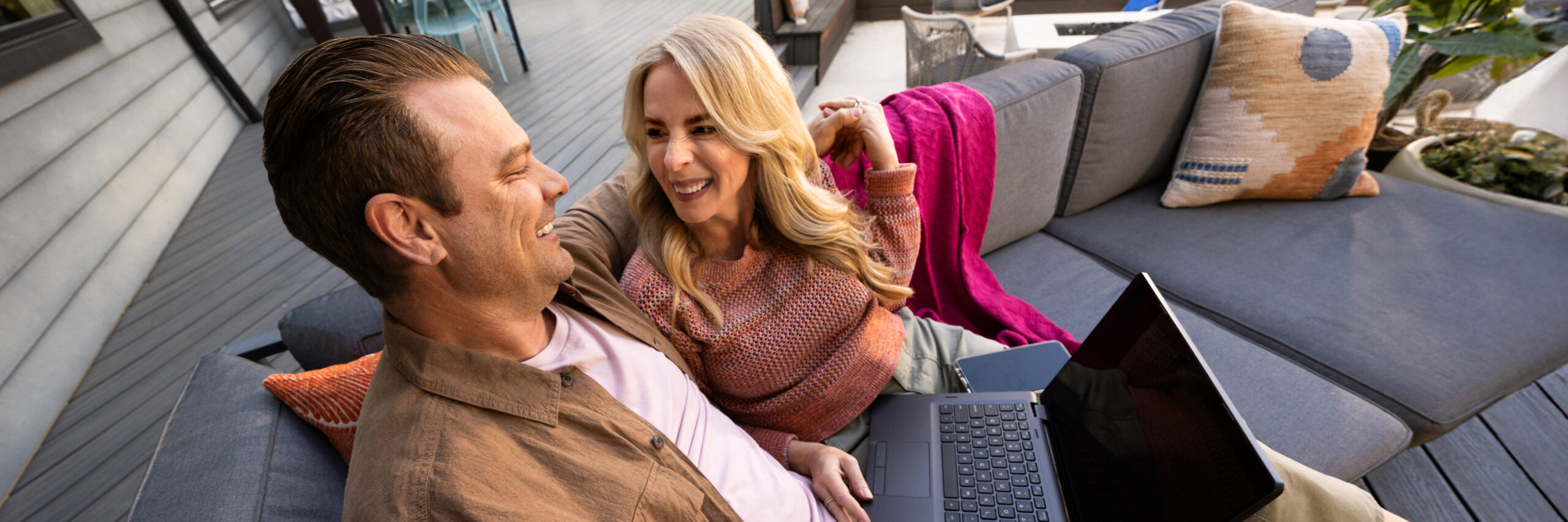 Two people sitting on a gray outdoor sofa, one holding a laptop, with a pink blanket and pillows.