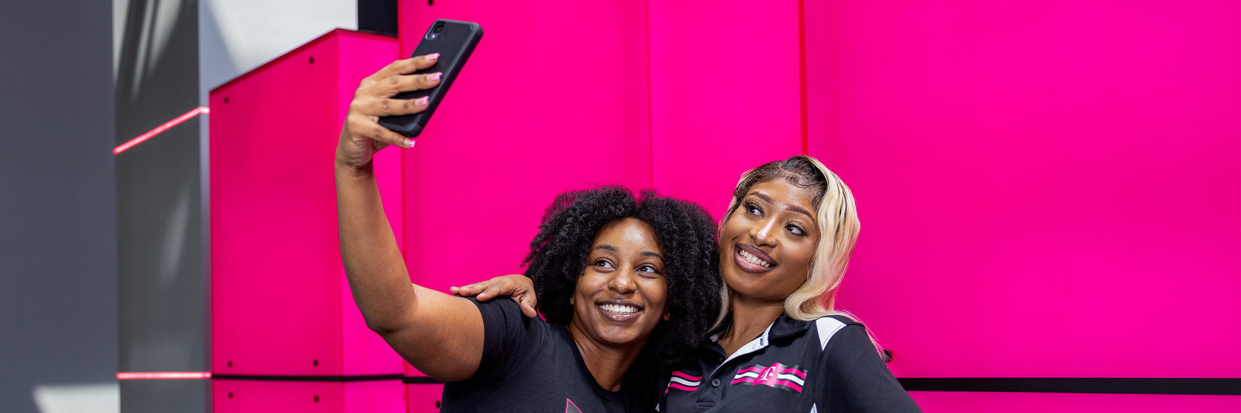 Two T-Mobile employees taking a selfie in front of a bright pink background.