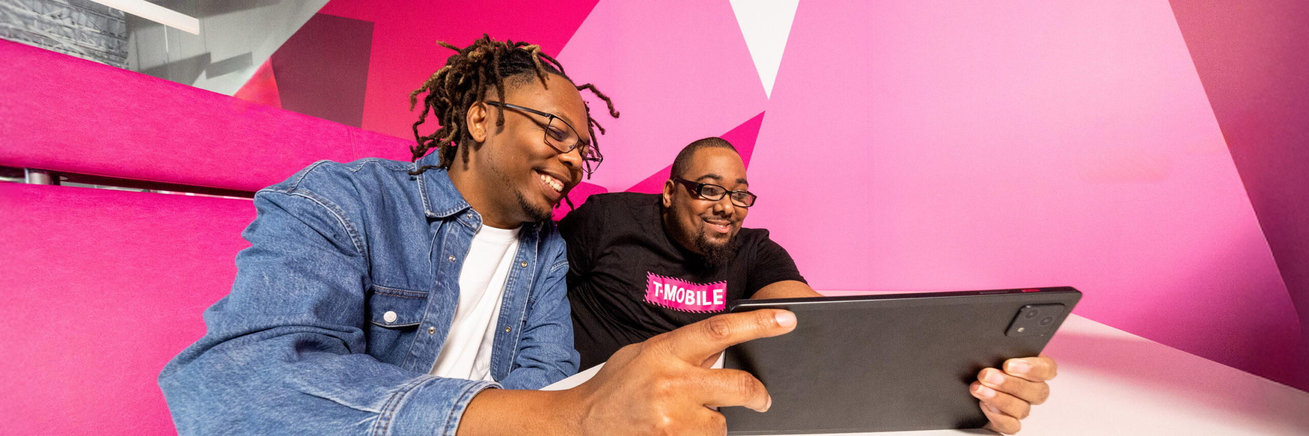 Two T-Mobile employees using a tablet on a pink bench with geometric background.