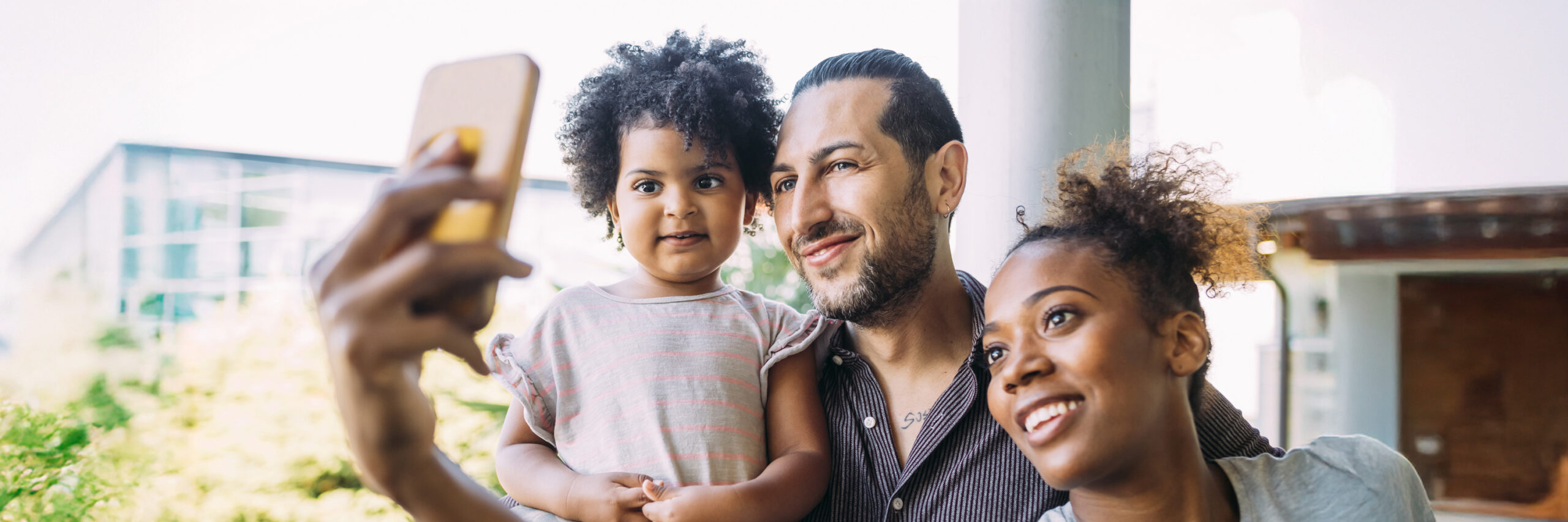 A family taking a selfie outdoors near greenery and a modern building.