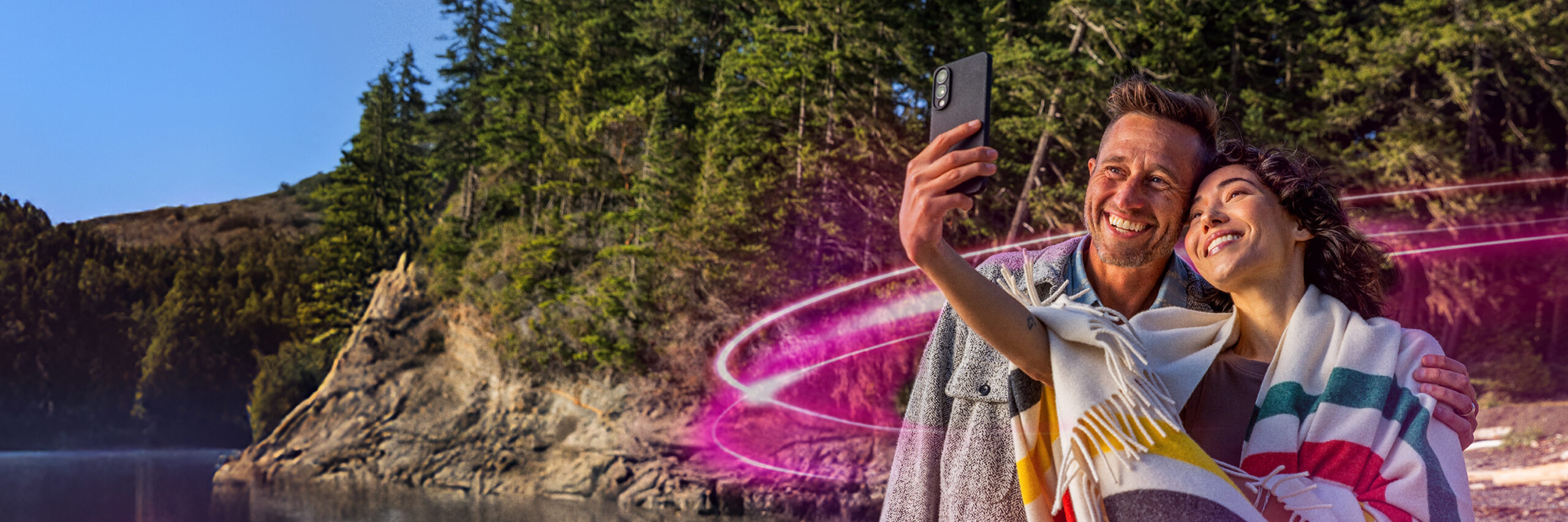 Two people taking a selfie near a rocky shoreline with trees and water, with a pink light trail effect.