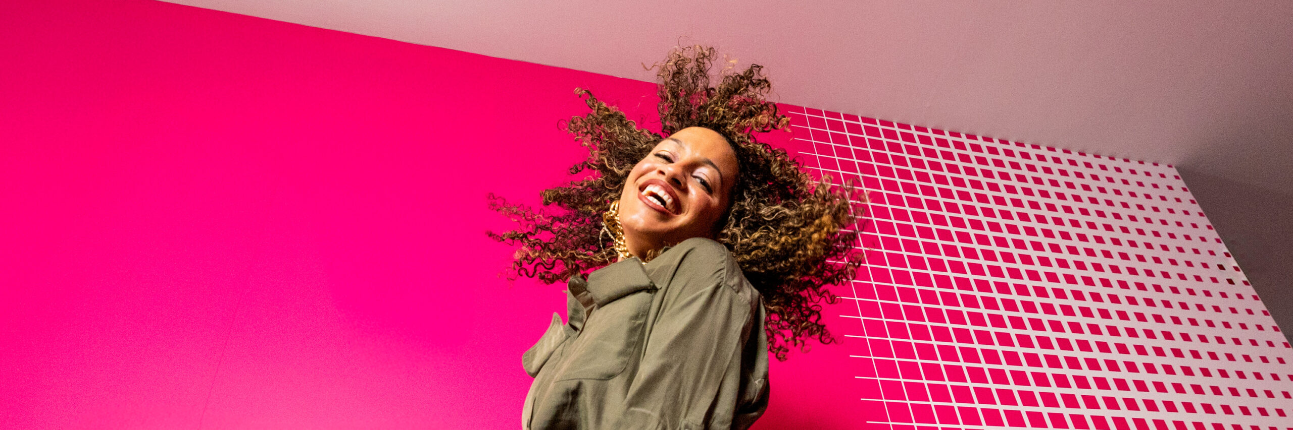A T-Mobile employee in a green jacket jumping against a pink background with white square patterns.