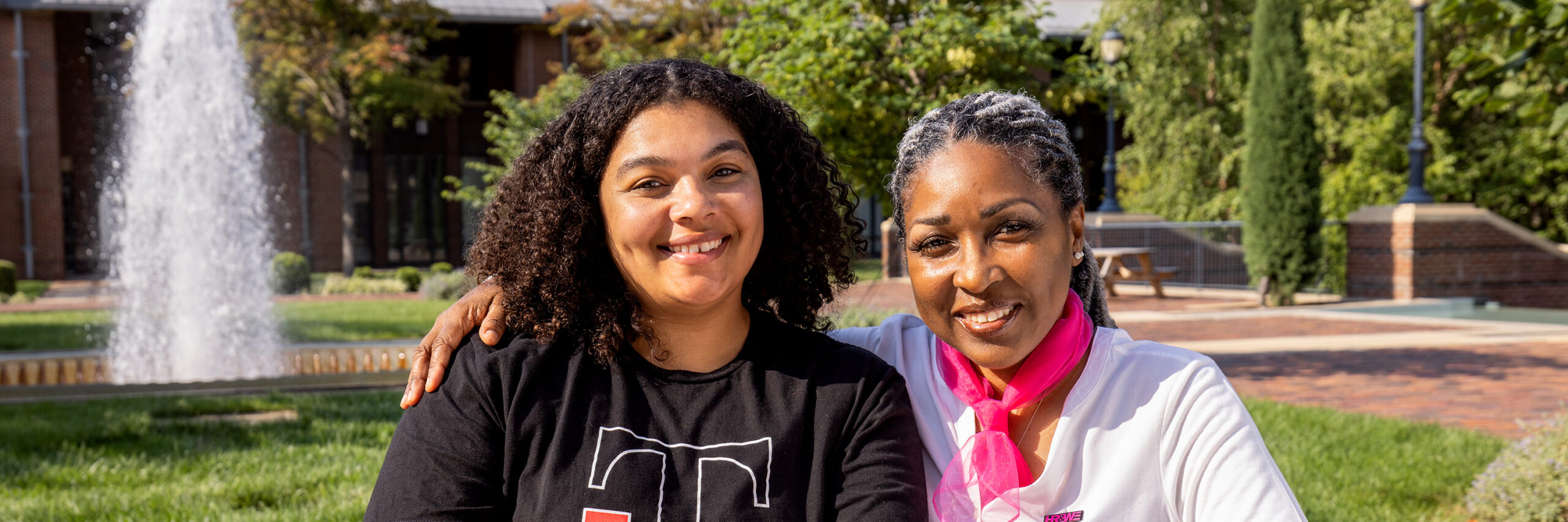 Two people sitting outdoors near a fountain with trees and buildings in the background.