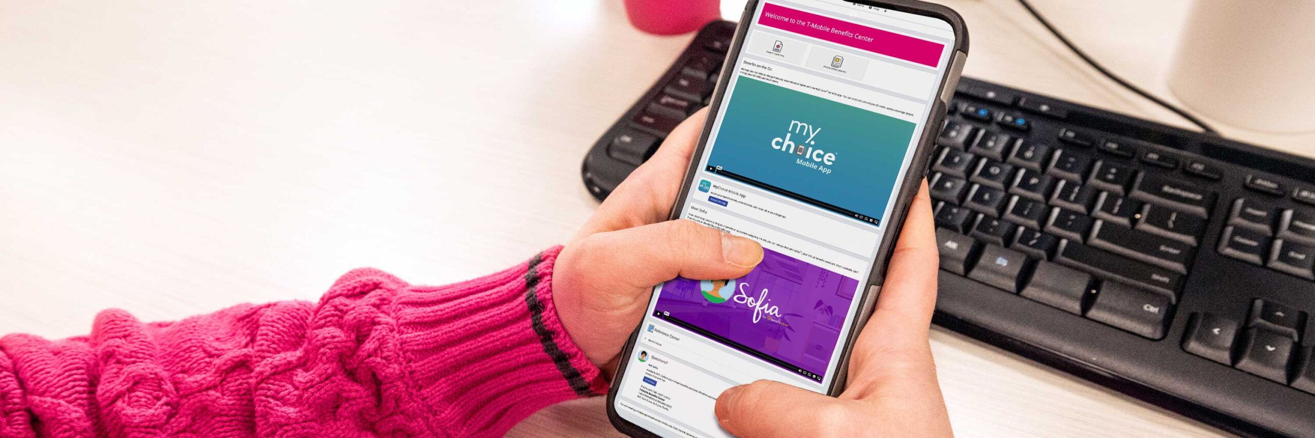 A person in a pink sweater holding a smartphone at a desk with a keyboard, and monitor.