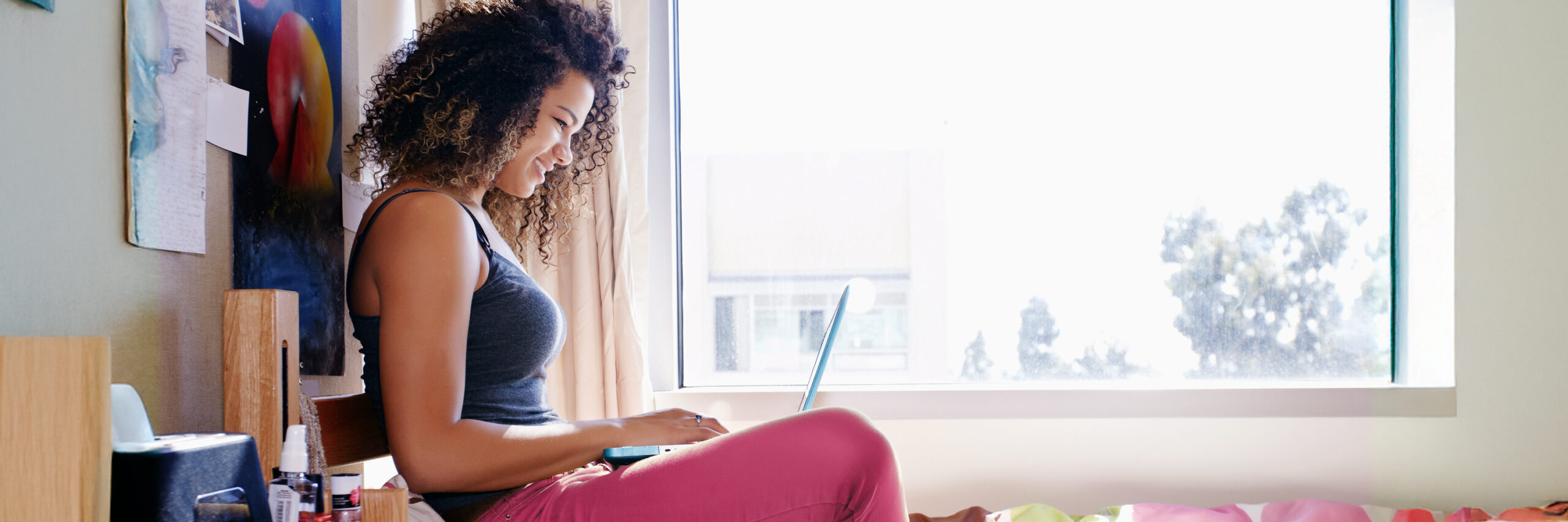 A person using a laptop on a bed near a window in a dorm room.