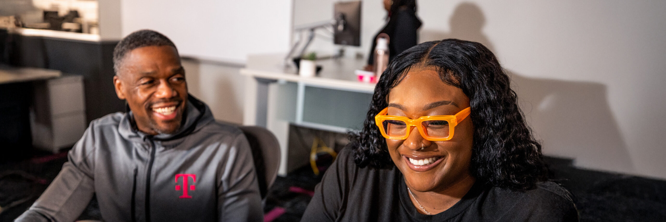 Two T-Mobile employees smiling and sitting in an office, with a desk and computer in the background.